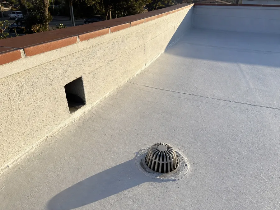 A coated low-slope roof meeting a stucco parapet, with a roof drain in the foreground