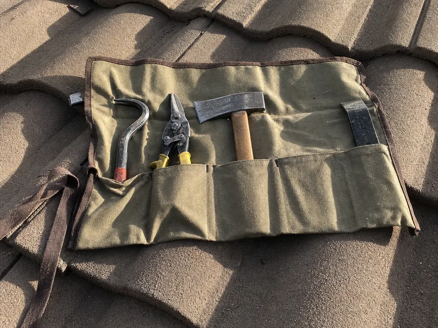 An open canvas tool roll on a tile roof with a shingle hatchet, tin snips and a pry bar in its pockets