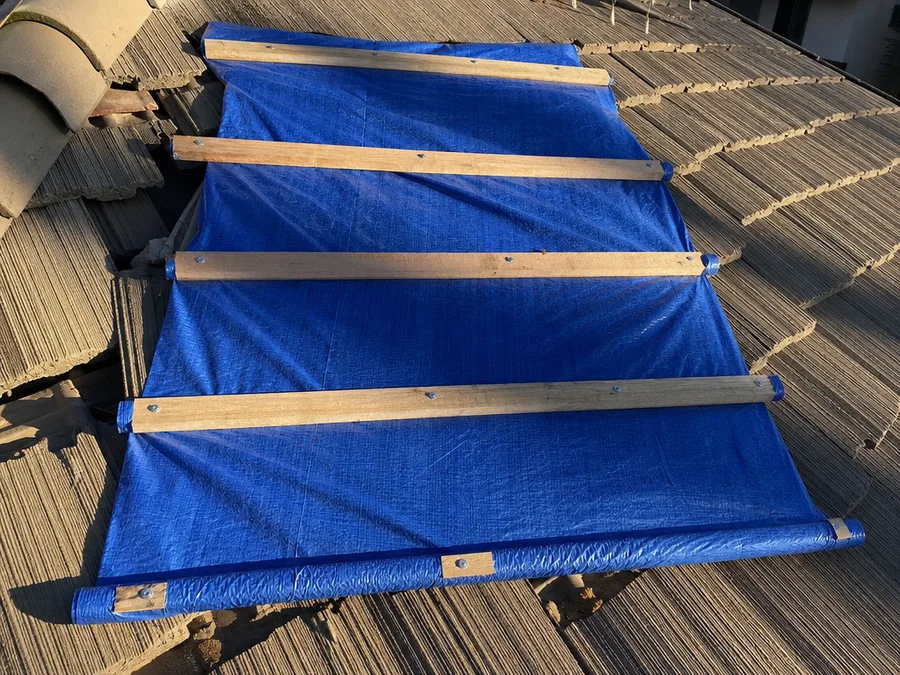 A blue tarp battened down over a damaged section of roof with timber strips screwed through it