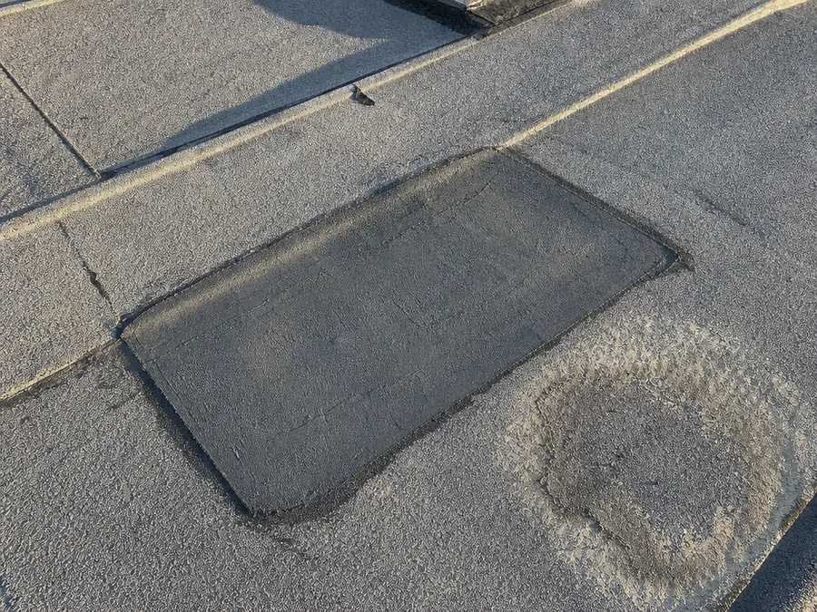 A repair patch bonded over a failed seam on a grey low slope roof, with a dried ponding ring beside it