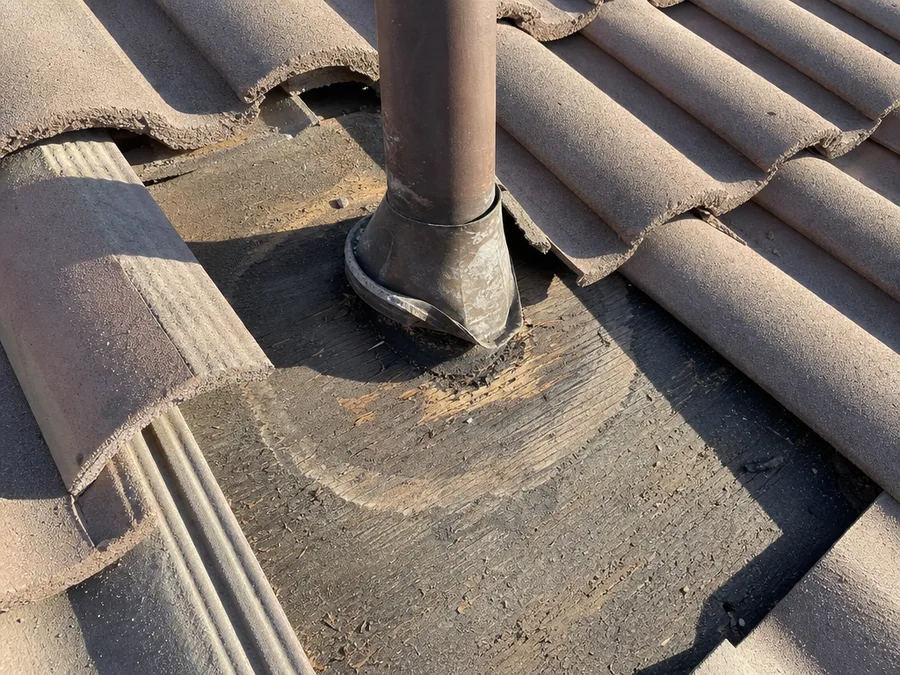 A plumbing vent flashing lifted away from a tile roof, showing dark water staining in the sheathing around the pipe
